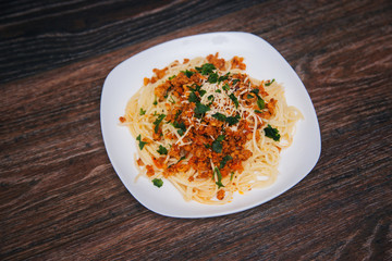 Spaghetti with minced meat in tomato with herbs and cheese on a white plate. Plate with food on a dark wooden table. Lunch with meat.