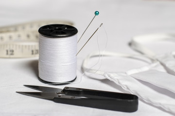 Needle and pin on white thread rolls for hand sewing on white cloth with scissors cut fabric and handmade mask close-up.