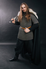 Warrior in chain mail and with a sword in a black cloak. Handsome guy with long hair and a beard in armor with weapons. Knight on a dark background. Studio photography