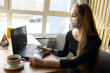 Beautiful girl sits and works at the table in a medical mask. On the table computer and tea .