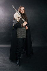 Warrior in chain mail and with a sword in a black cloak. Handsome guy with long hair and a beard in armor with weapons. Knight on a dark background. Studio photography
