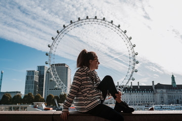 Young woman sitting on a wall in London with a Ferris wheel in the background wearing a black and white striped coat. She is wearing a ponytail, black pants, black sneakers and glasses.