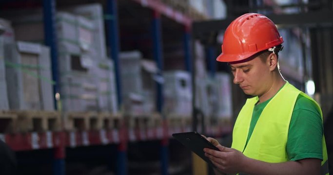 An engineer examines a goods in a warehouse and notes on a tablet