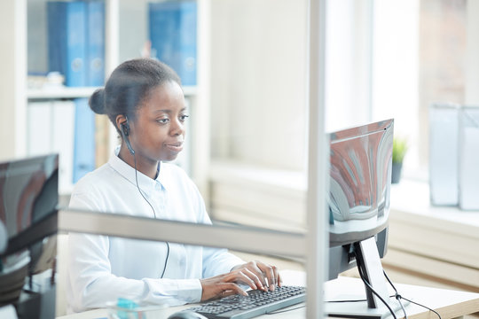 Portrait Of African-American Businesswoman Wearing Headset While Using Computer At Workplace, Shot From Behind Glass Wall, Copy Space