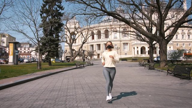 Beautiful young and fit woman running and jogging alone in city center wearing protective face mask, slow motion