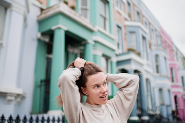 Attractive natural young girl with blue eyes, blonde hair and nude sweater, combing her hair on a street with colorful houses in London.