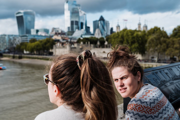 Couple of women in fashion clothes, leaning against the wall of a bridge while one of them observes the river and the city of London and the other looks at the camera. Both of them enjoy a cloudy autu