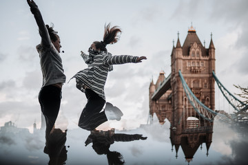 Two girls laughing and jumping in front of Tower Bridge, London. Cloudy day with a reflection of the lower half of the photo.