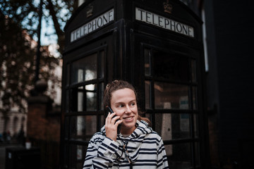 Portrait of a young woman dressed in white autumn clothes with black stripes and talking on the phone, while waiting in front of a typical black London telephone box where she can read "TELEPHONE".
