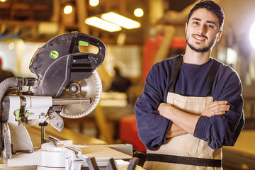 portrait of young caucasian positive carpenter at work place, handsome guy in work clothes smile and look at camera, enjoy being woodworker
