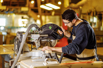 confident young carpenter work with mitre saw in factory, stand standing behind a workbench wearing uniform. industry, handicraft, carpentry concept