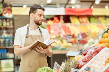 Waist up portrait of bearded farmer holding notebook while selling fresh fruits and vegetables at market stand, copy space