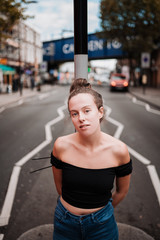 Portrait of a young woman dressed in trendy clothes and with her hair up, leaning against a lamppost in the middle of a busy London street.