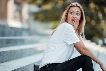 Beautiful blonde girl sit with long hair flying on the wind, make different face expressions. Woman looking at camera outdoors wearing fashionable white t-shirt.