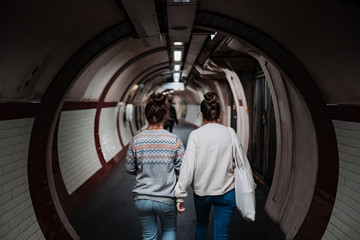 A modern female couple walks down a long dimly lit corridor inside a London underground station. Both are photographed from behind.
