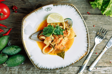 Restaurant dish on a wooden background with vegetables. Soup with fish and seafood and lemon on a plate.