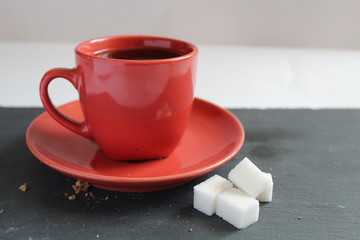 tea or coffee in a red Cup and saucer stands on a gray stone or Board table and there are pieces of sugar cubes next to it