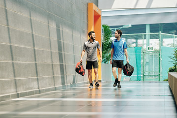 Handsome young sportsmen with gym bags walking along health club corridor and discussing news