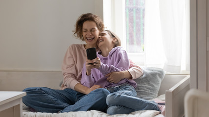 Naklejka na ściany i meble Cheerful mom and teen daughter laughing using smartphone app at home sitting on bed. Happy positive family adult parent having fun watching funny videos enjoying lifestyle relaxing with teenage girl.