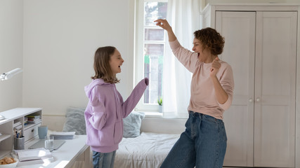 Happy family young mom and teenage daughter having fun dancing at home together. Cheerful mother with cute adolescent teen girl laughing, singing, enjoying funny weekend lifestyle activity in bedroom.