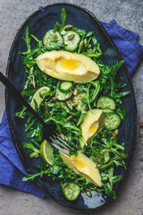 Green salad with cucumber, avocado and arugula in blue dish, dark background. top view.