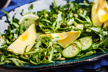 Green salad with cucumber, avocado and arugula in blue dish, dark background.