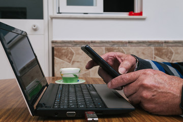 Older man typing laptop and operating mobile phone at home. Telecommuting during confinement. Communication with family members during quarantine. Selective focus.