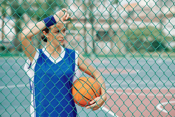 Female basketball player leaning on wire mesh fence around outdoor court and looking away