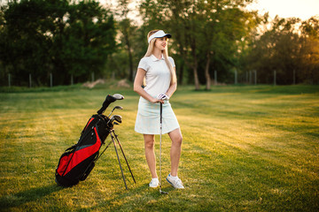 Fototapeta premium An attractive sports girl golfer is standing on the golf course near equipment. Waiting for a good game of golf. Golf Club.