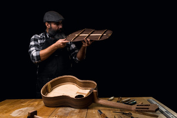 Luthier wearing cap, plaid shirt and apron. Measuring back of Spanish guitar. On the workbench guitar in construction and tools. Dark black background. - obrazy, fototapety, plakaty