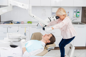 Fototapeta premium Adorable little girl performing an examination of a female doctor, the doctor is lying in a dentist chair. Dentist and child in a dentist office. Little girl playing in the dentist's office
