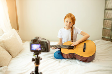 portrait of beautiful cute caucasian child girl blogger playing guitar at camera. adorable girl sit showing, performing new melody for subscribers. vlog, blog, video concept
