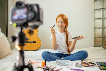 Fototapeta na wymiar portrait of little caucasian blogger child interestingly talk about cosmetics, cute girl with long red hair present beauty things, sit in front of camera. broadcast