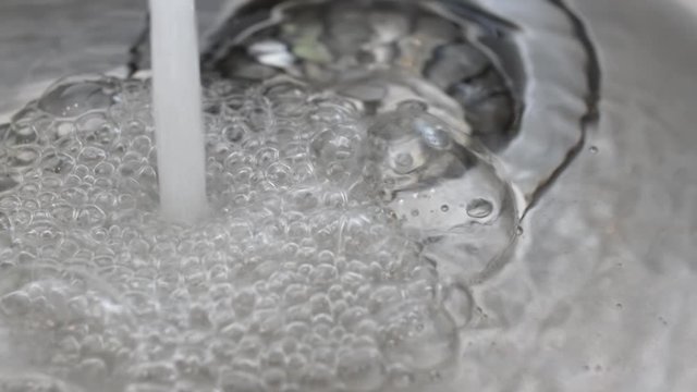 Water flows from a faucet to a sink full of water. Air bubbles in the water. In slow motion.