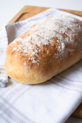 A loaf of homemade fresh bread with flour sprinkled on top in a kitchen towel on wooden cutting board with slices