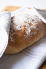A loaf of homemade fresh bread with flour sprinkled on top in a kitchen towel on wooden cutting board with slices