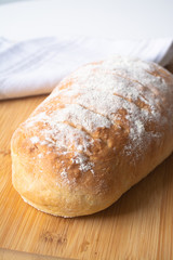 A loaf of homemade fresh bread with flour sprinkled on top in a kitchen towel on wooden cutting board with slices