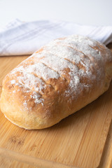 A loaf of homemade fresh bread with flour sprinkled on top in a kitchen towel on wooden cutting board with slices