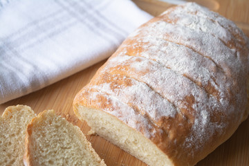 A loaf of homemade fresh bread with flour sprinkled on top in a kitchen towel on wooden cutting board with slices