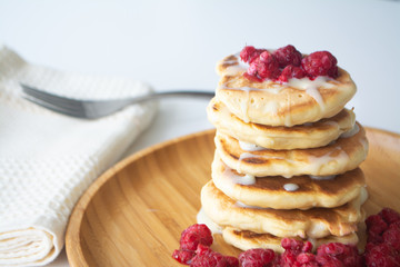 Fluffy pancakes with condensed milk and raspberries on a wooden plate with a kitchen napkin and a fork 
