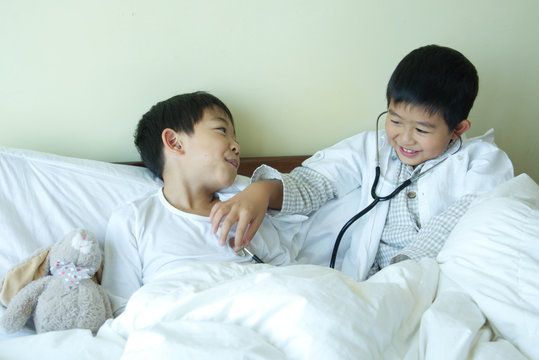 Two cute Asian boys are playing a doctor and patient play role on the bed at home with fun and smile.One kid is with stethoscope on.Learning development and career imagination of children concept.