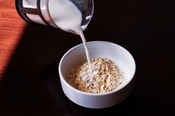 Pouring the milk into oatmeal in a white porcelain bowl on the wooden background.