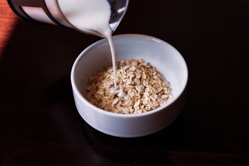 Pouring the milk into oatmeal in a white porcelain bowl on the wooden background.