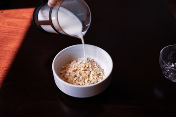 Pouring the milk into oatmeal in a white porcelain bowl on the wooden background.