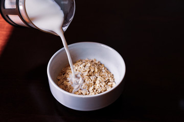 Pouring the milk into oatmeal in a white porcelain bowl on the wooden background.