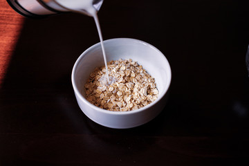 Pouring the milk into oatmeal in a white porcelain bowl on the wooden background.