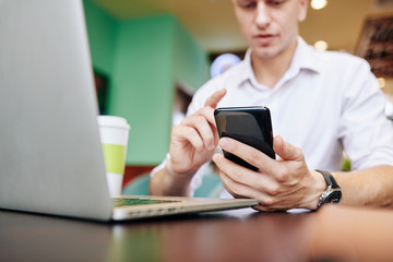 Young businessman sitting at desk with opened laptop and checking notifications and messages in his phone