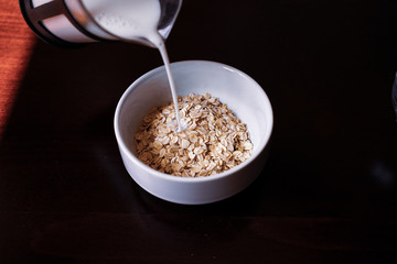 Pouring the milk into oatmeal in a white porcelain bowl on the wooden background.
