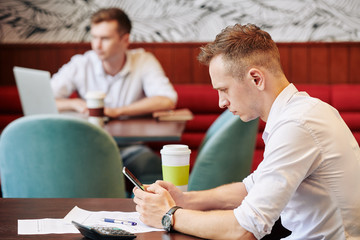 Serious frowning young businessman sitting at cafe table with documents in front of him and reading news on smartphone screen
