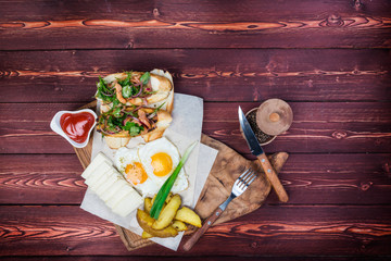 Big breakfast. Bruschettas, fried eggs, fried potatoes and cheese with sauce. There is a black pepper and cutlery. Top view on a wooden background